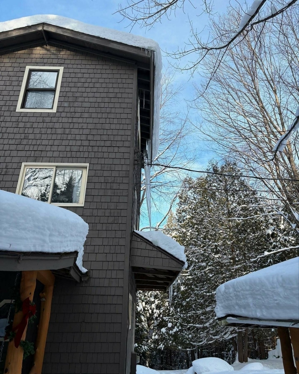 Icicles hang from a snow-covered roof of a dark wooden house nestled in a wintery landscape with bare trees and evergreens under a blue sky.