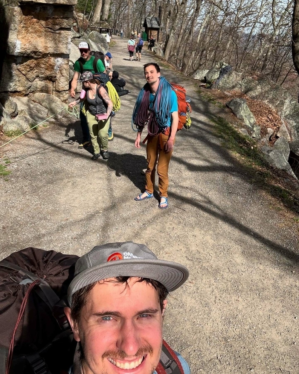 People carry climbing gear on a forested trail smiling and walking. A leafless wooded area with rocks and a stone structure surrounds them. Person in foreground wears a cap with &ldquo;ONE MOVEMENT&rdquo;.