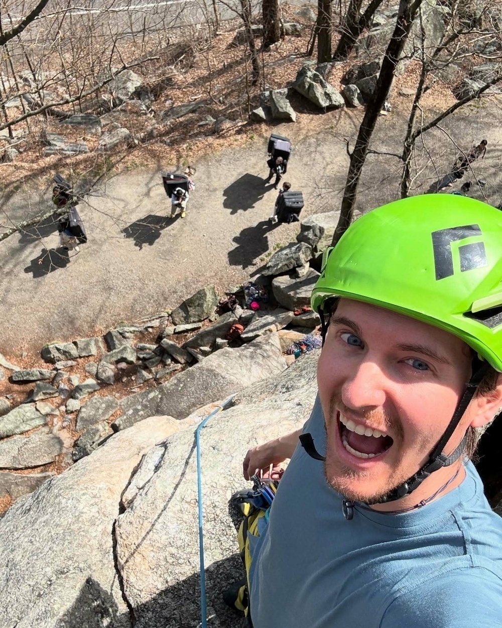 Man wearing a green helmet smiles while standing on a rocky cliff edge holding a climbing rope in a wooded area with people below carrying gear.