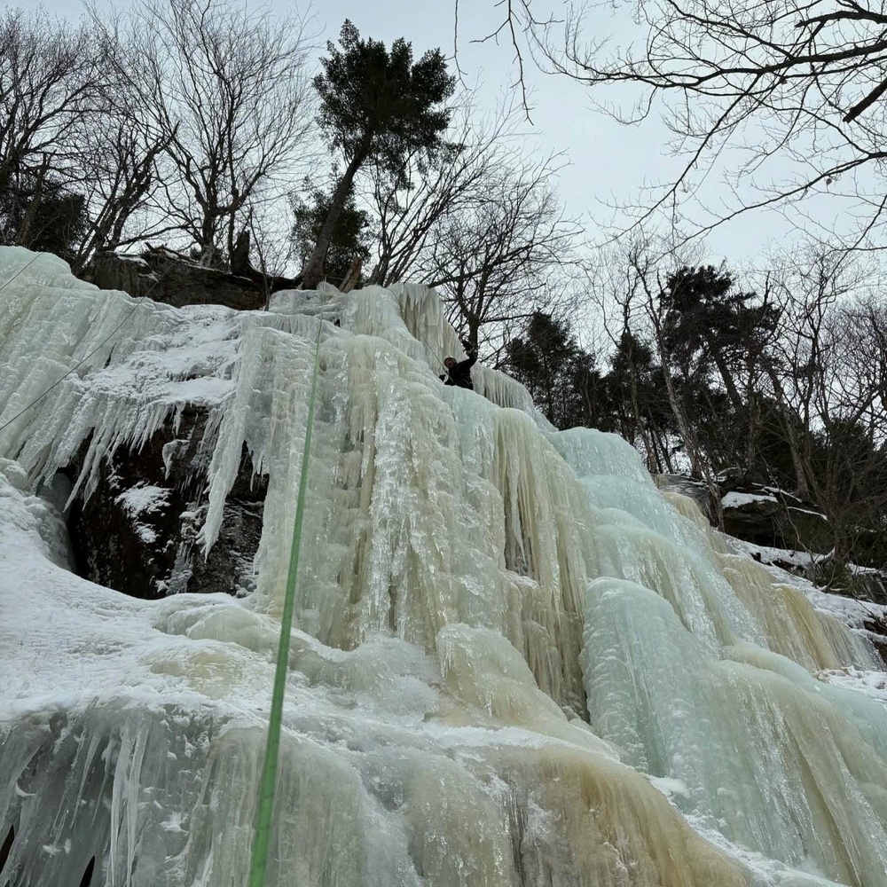 A climber ascends a large frozen waterfall using climbing gear surrounded by tall trees and a cloudy sky.