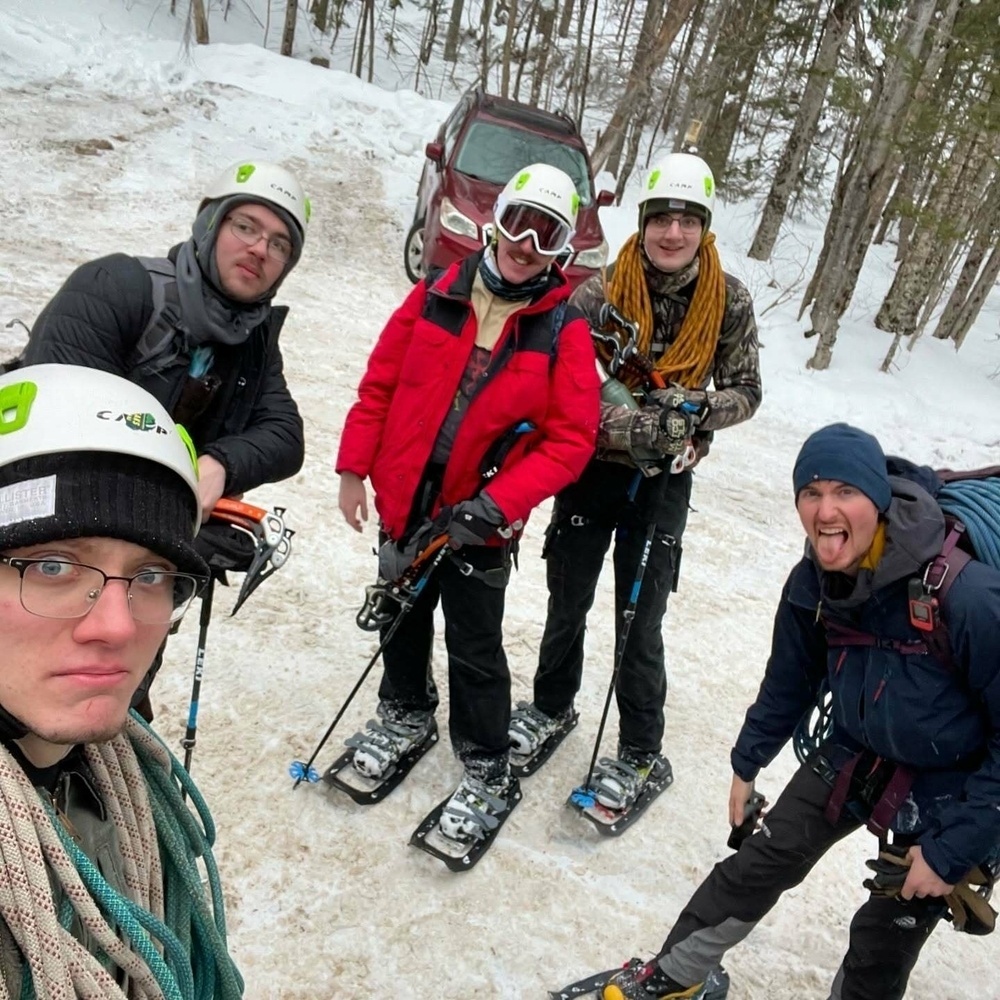 Five people in snow gear smile and pose on a snowy path wearing helmets and snowshoes. One person is playfully sticking out their tongue. Trees and a parked car are in the background.