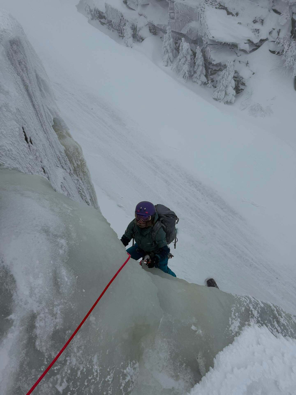 Climber ascends an ice-covered cliff using a red rope for support thick snow blankets the surrounding rocky landscape and evergreen trees creating a wintry alpine environment.