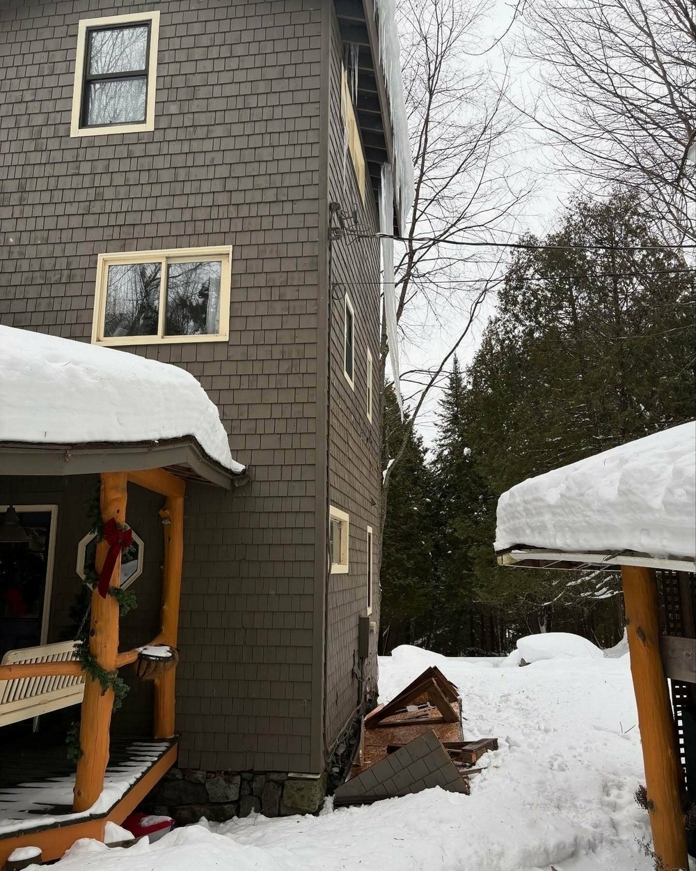 Tree-covered house with decorative porch and fallen roof section surrounded by heavy snow, set in a wooded area.