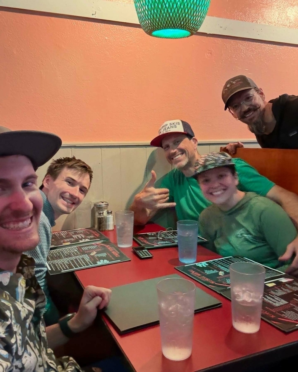 Five people smiling and posing at a restaurant table holding menus. A green lamp hangs above, creating a warm, relaxed atmosphere. One person wears a hat with &ldquo;Two Skis Means&rdquo; text.