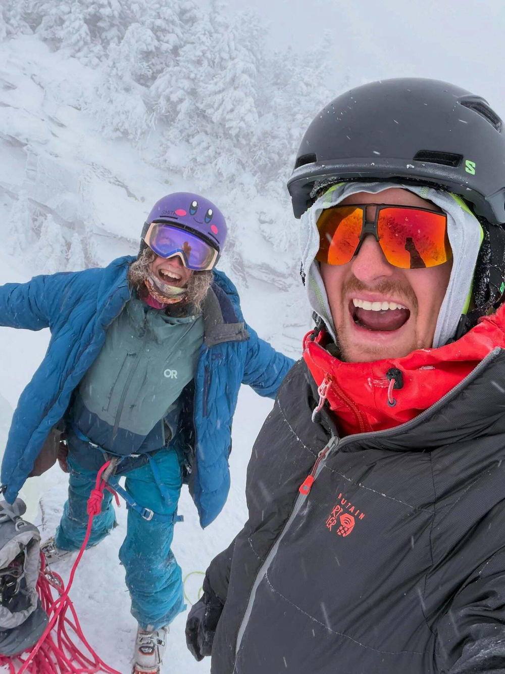 Two people wearing winter gear smile at the camera amidst a snowy landscape. One person wears a blue jacket with OR logo and ski goggles. The other wears a black helmet and sunglasses.