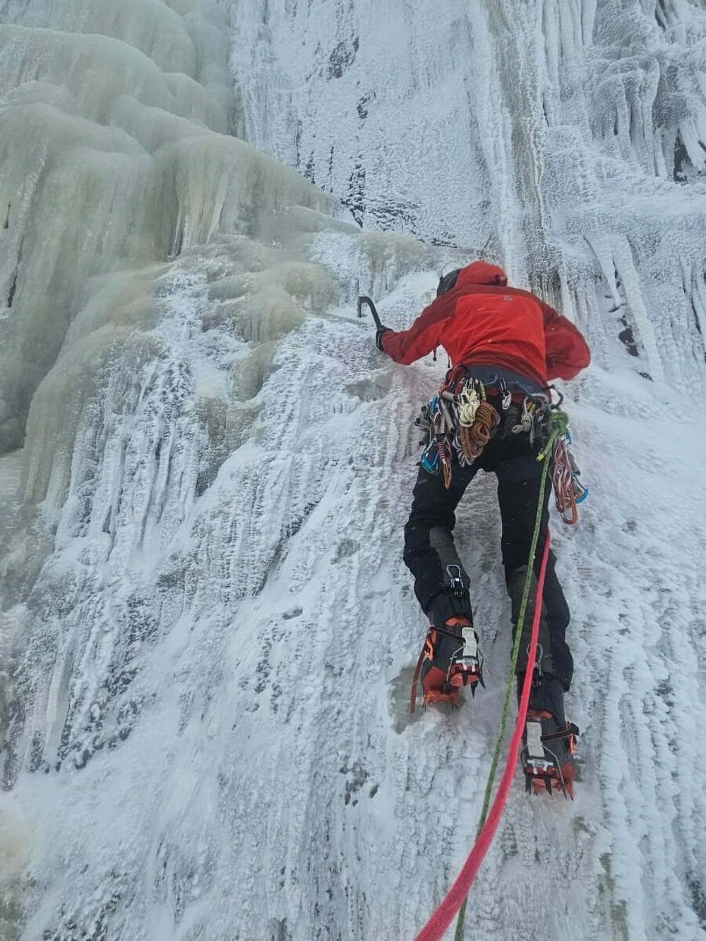 A climber ascends a steep icy wall using ice axes and crampons. They wear a vibrant red jacket, surrounded by rugged ice formations.