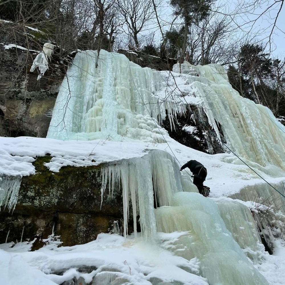 A person climbs a large frozen waterfall, surrounded by snow-covered rocks and trees in a wintry landscape.