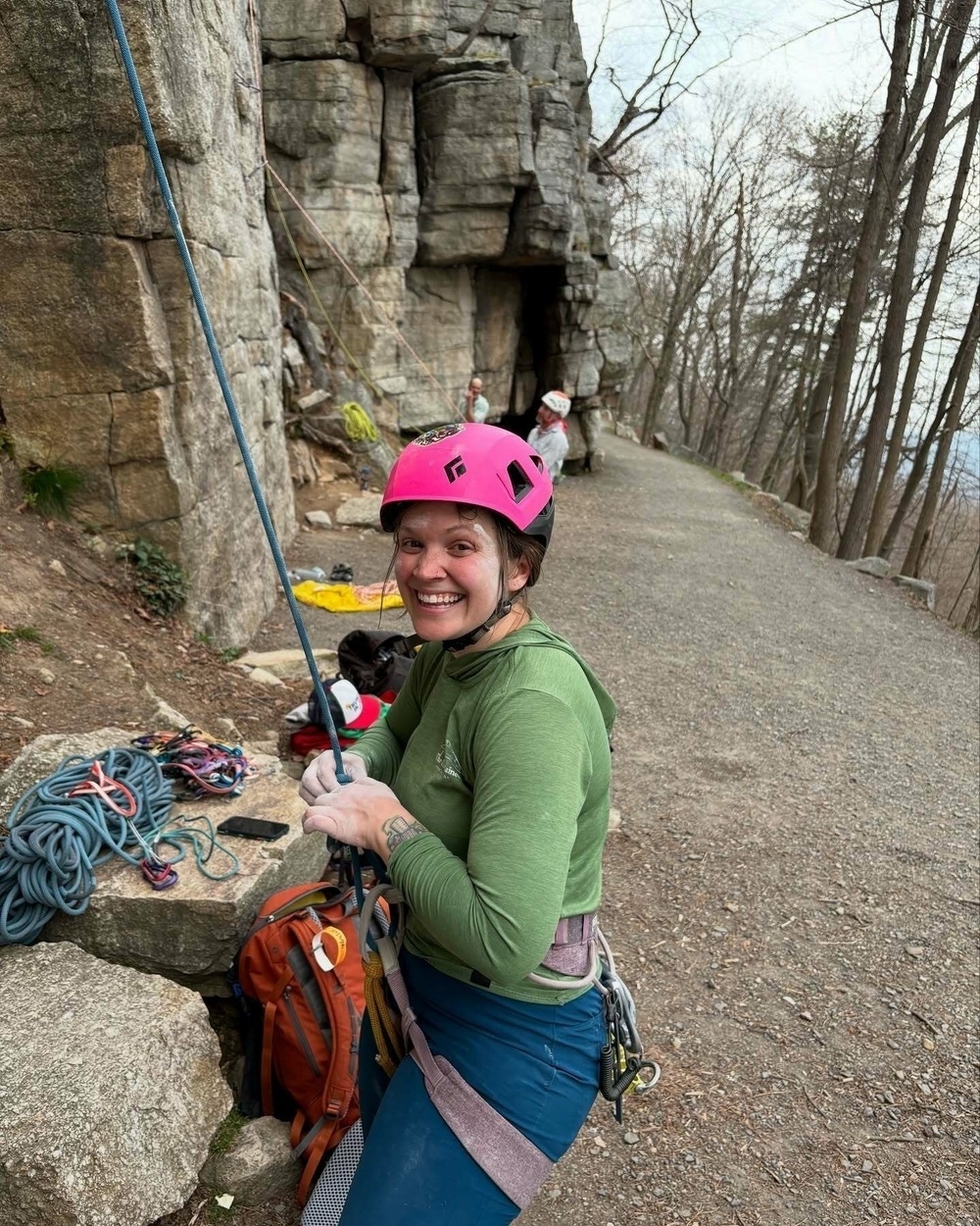 A climber wearing a pink helmet holds a rope and smiles while gear is arranged beside them. They are near a rocky cliff in a wooded outdoor setting.