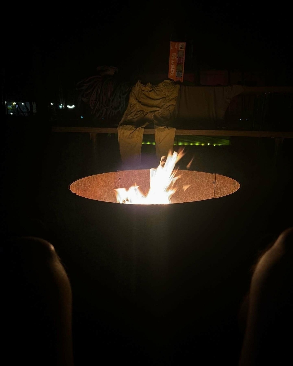 A fire pit burns with bright flames at night on a dark surface with pants and a box labeled Sam&rsquo;s Choice on a bench nearby in the background.