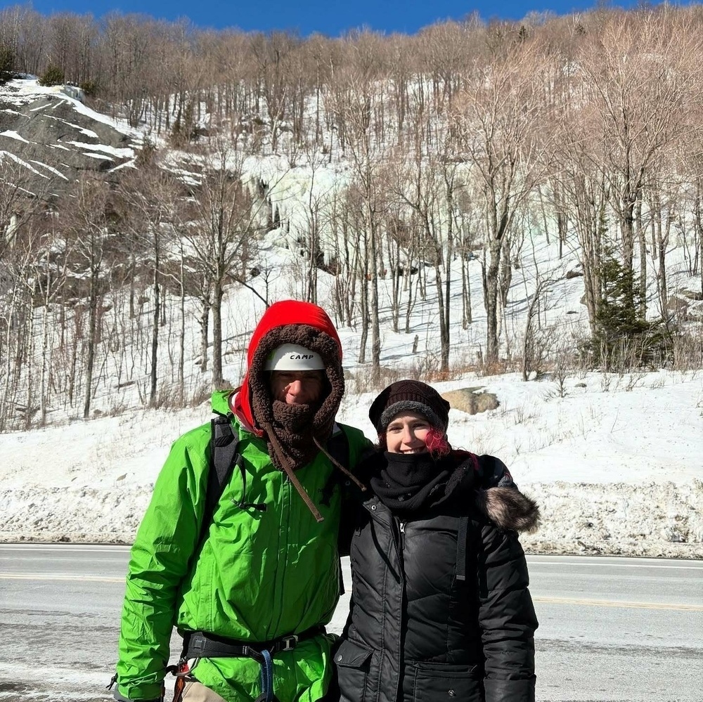 Two people bundled in winter clothing stand closely in front of a snowy mountain hillside with leafless trees under a clear blue sky.