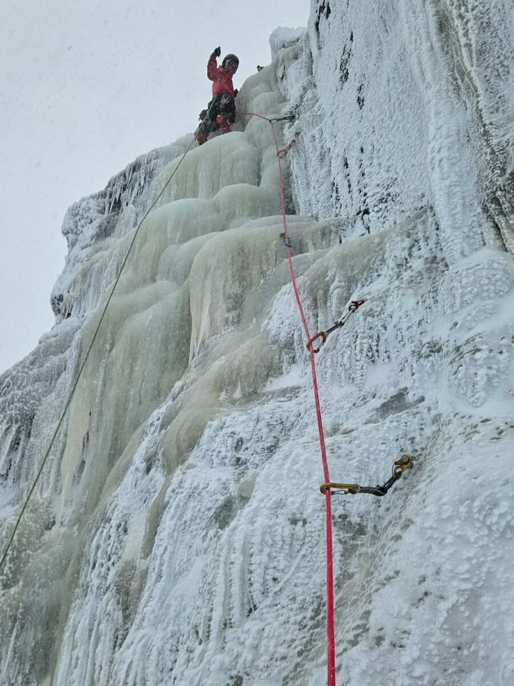 A climber is ascending an icy cliff using ropes and ice-climbing gear in a snowy mountainous environment. The climber is wearing a red jacket and is secured with a red rope.