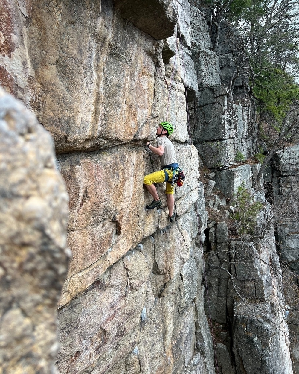 A climber wearing a helmet and harness ascends a rocky cliff face surrounded by rugged terrain and scattered trees.