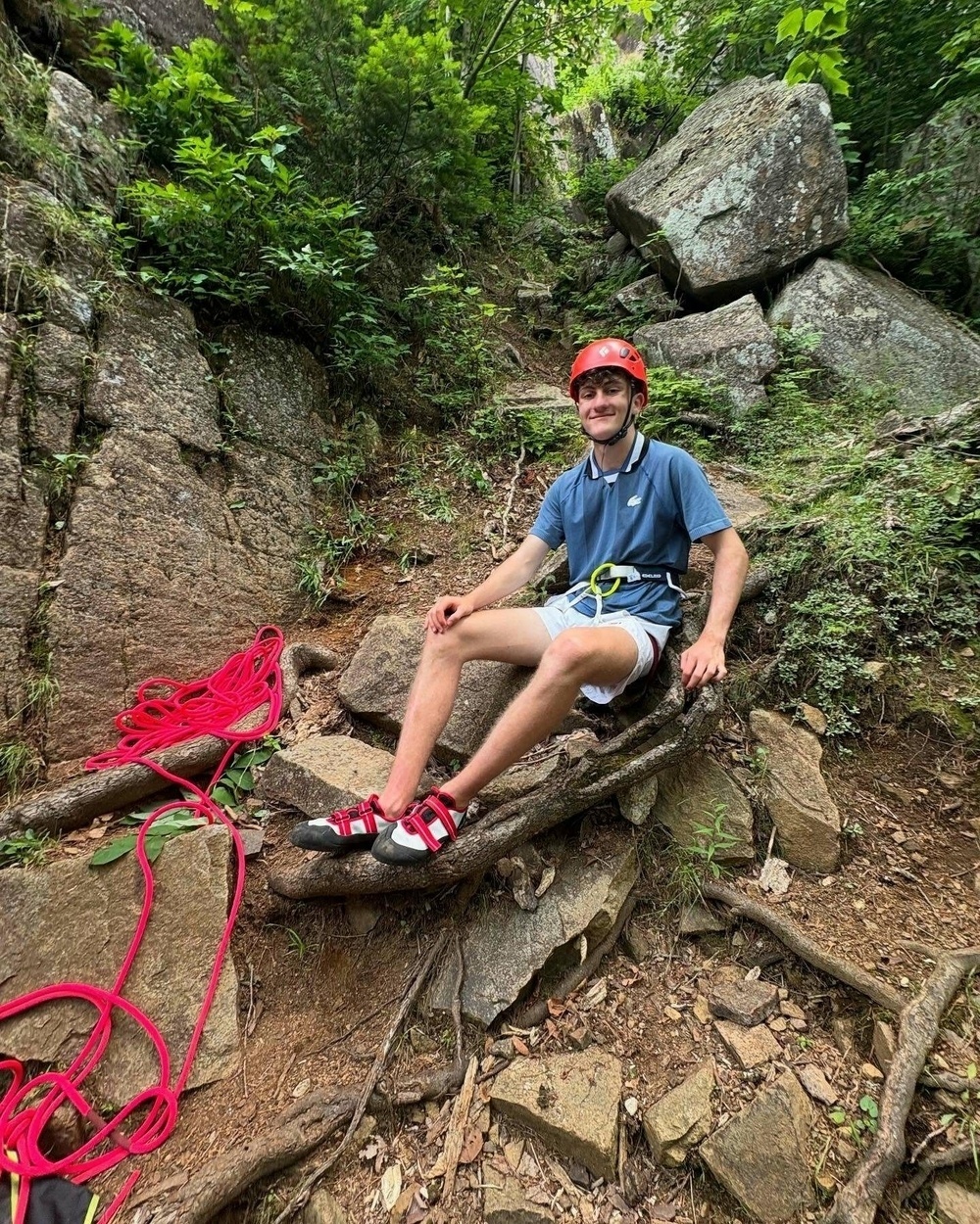 A person is sitting on a rock wearing a red helmet and climbing gear smiling in a forested rocky area with a coil of red rope beside them.