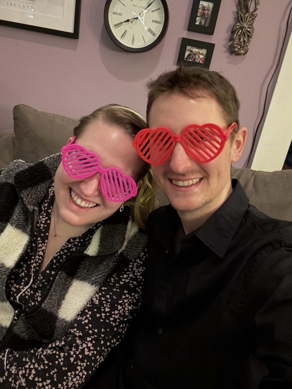Two people wearing heart-shaped novelty glasses smile while sitting on a couch. A clock and framed photos hang on the purple wall behind them.