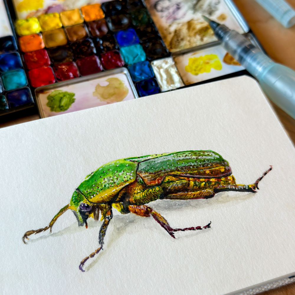 A detailed watercolor painting of a vibrant green beetle on a piece of watercolor paper, with a palette of colorful paints and a water brush beside it.