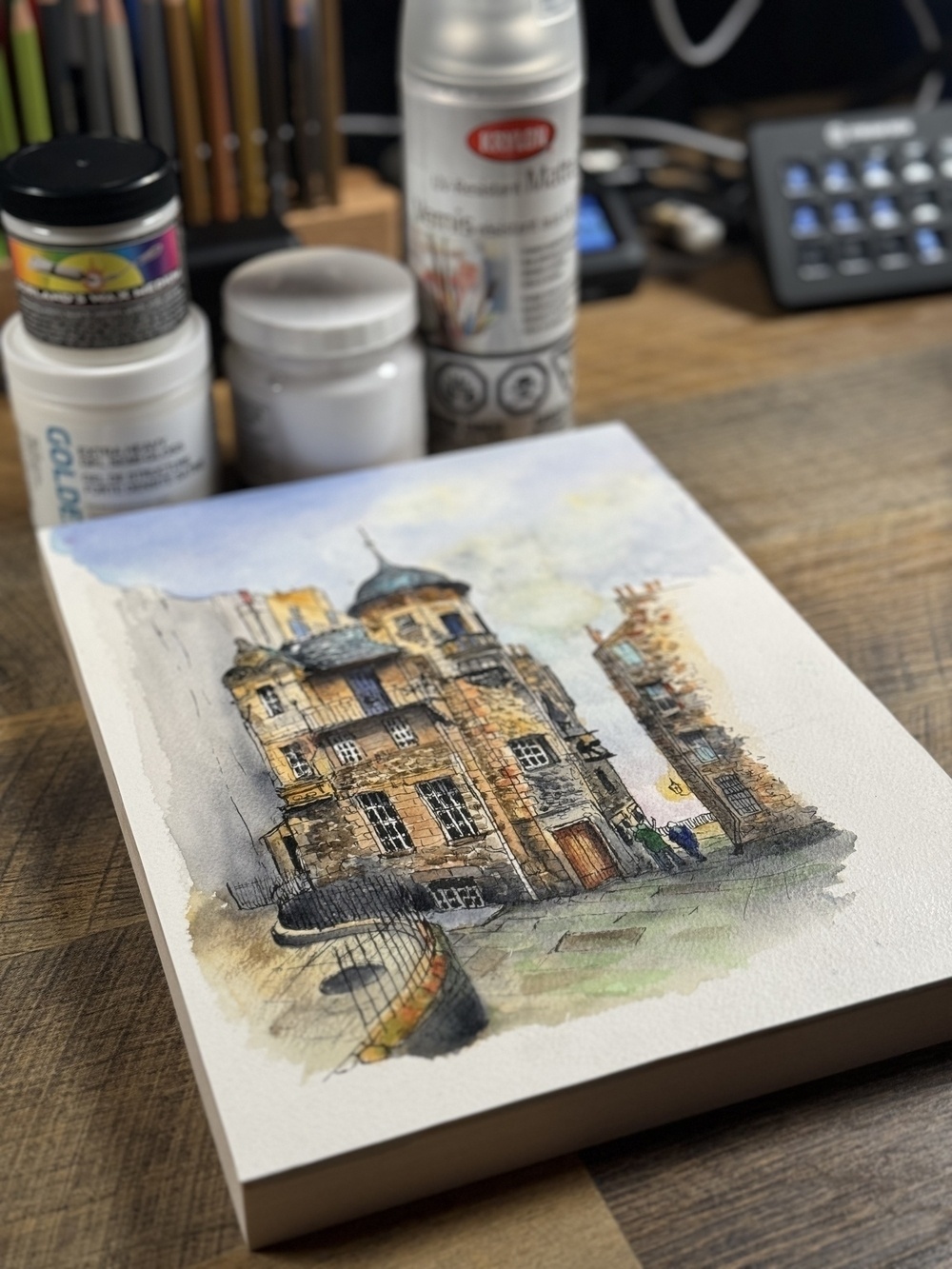 A watercolor painting of a historic building sits on a wooden table surrounded by art supplies like paint bottles and spray cans.