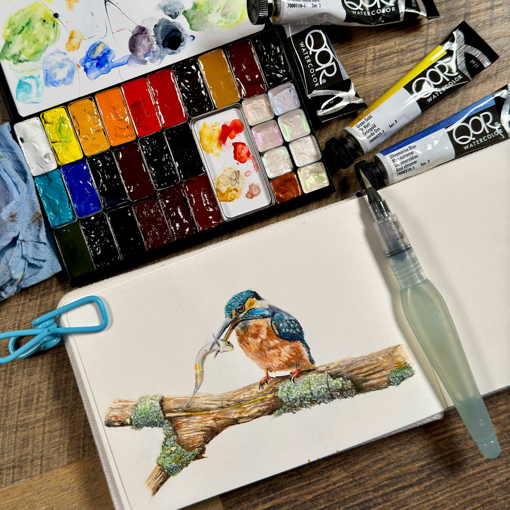 Watercolor painting of a common kingfisher holding a small fish in its beak, perched on a mossy branch. The artwork is surrounded by a QoR watercolor palette, paint tubes, and a water brush on a coffee shop table. 