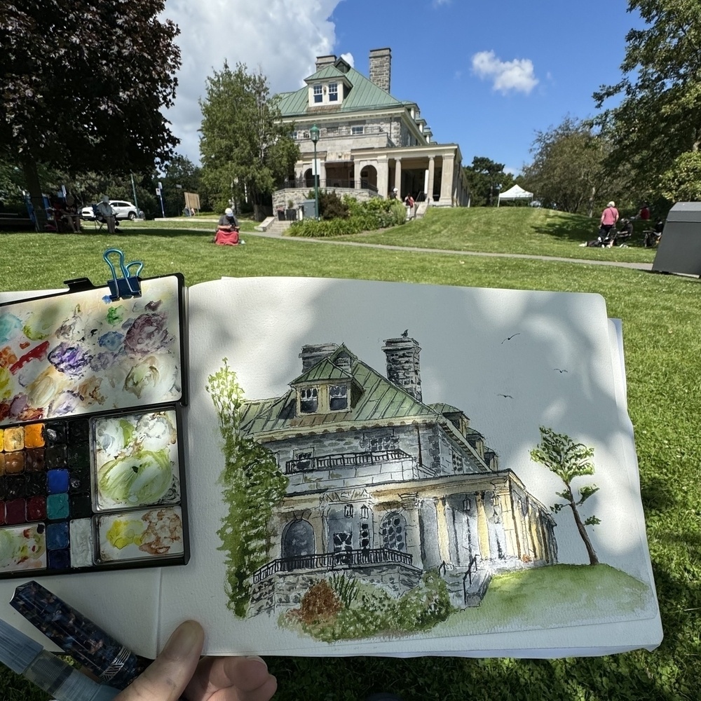 A person is painting a watercolor of a historic house in a park, with the actual house visible in the background.