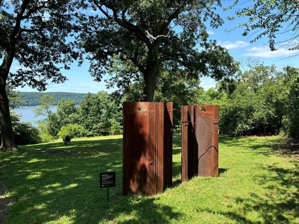 A rusty, brown metal sculpture stands in a grassy park surrounded by trees and overlooking a body of water with a small sign nearby.