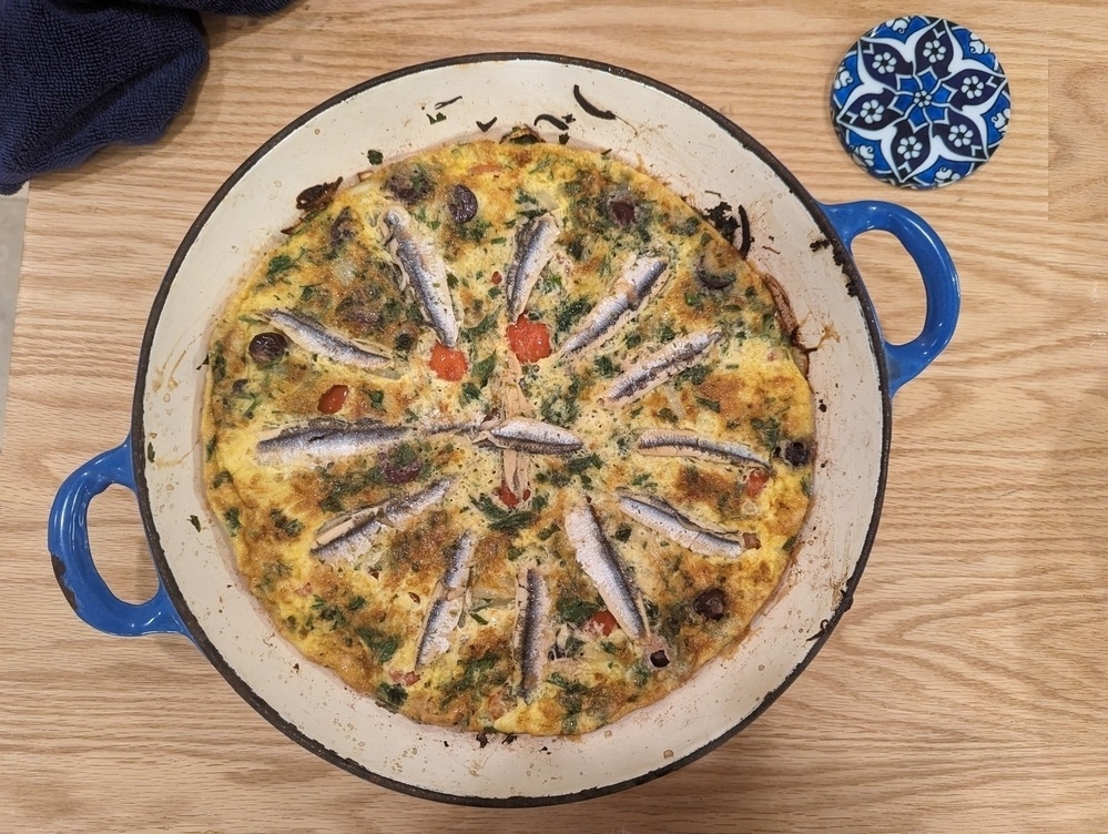 A frittata with tomatoes, olives and anchovies, in a blue pan, on a wooden surface.