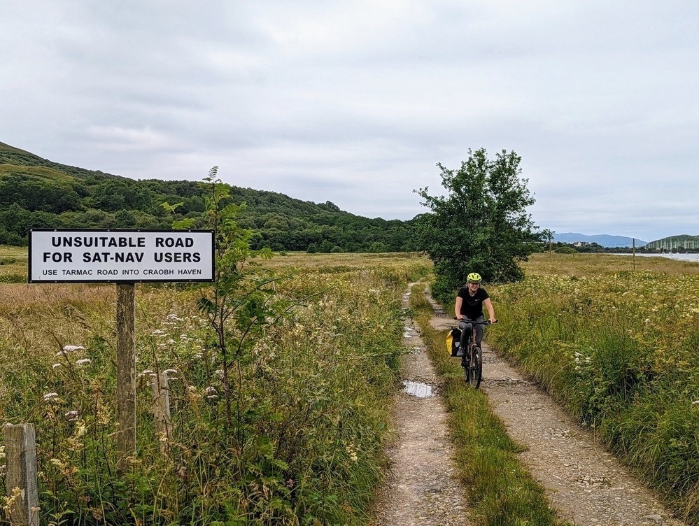 A cyclist on a deeply-rutted rural farm track, approaching a large sign that reads: Unsuitable road for Sat-Nav users.