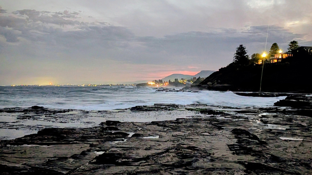 A rocky shoreline extends into a foamy sea under a cloudy sky, with distant city lights and a few illuminated houses perched on a hill.