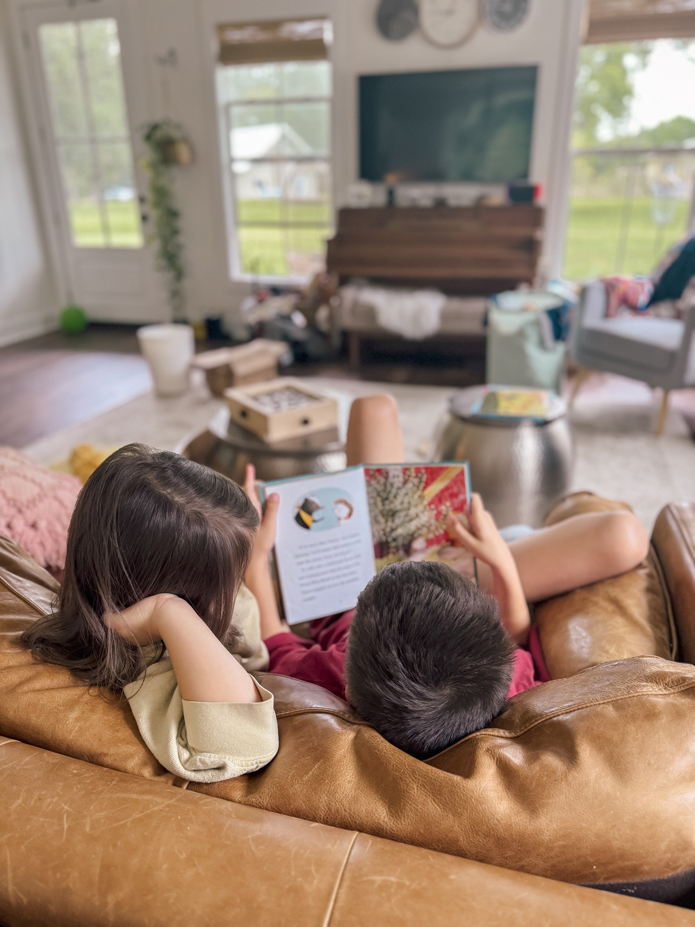 A photo from behind of a brother reading a book to his little sister while both are snuggled on the sofa 