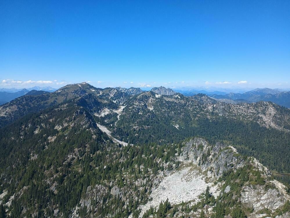 A ridge leads away from the foreground to the left, rising and falling between peaks before running into a range of jagged mountains in the middle distance. Past that, a crowd of mountains marches off to the horizon. In the very center of the far distance, a snowy stratovolcano rises above them all.