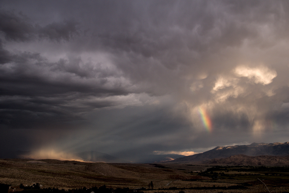 A large valley is bordered by mountains to the south and east. Over the southern mountains there are storm clouds and a rainbow.  The setting sun pierces a gap in the storm and lights up the mountains to the east, the rays visible where scattered by the rain.