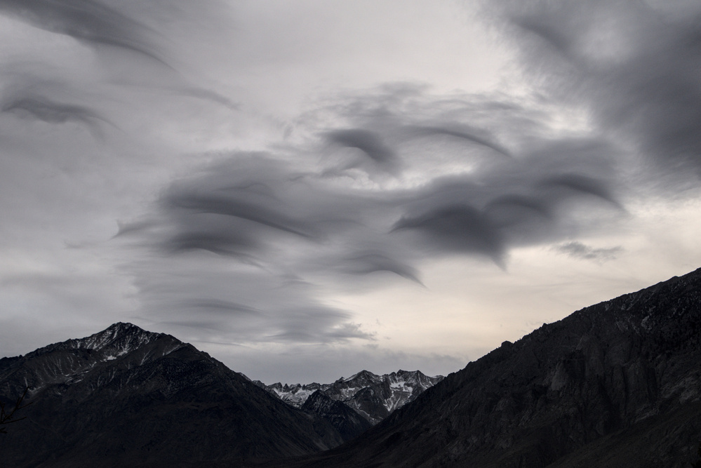 A collection of wispy, curved, grey clouds hangs over a mountain landscape. 
 The clouds have some darker, curved streaks that look vaguely like dolphins swimming.