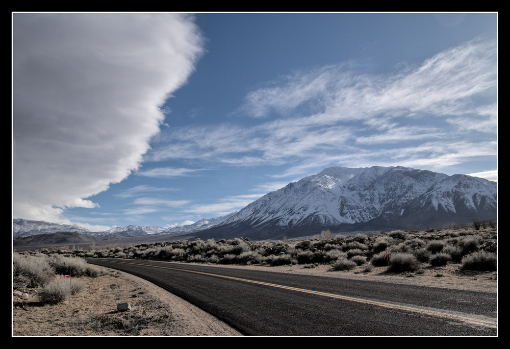 A road curves to the left, while above, a huge cloud curves upward, balancing the road.  There is blue sky to the right of the cloud, leading to a snow-covered granite mountain.