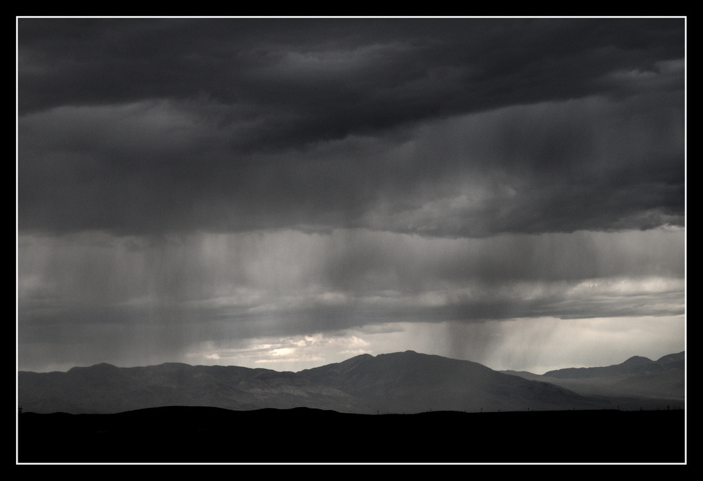 Dark clouds hover over a mountain range. Streaks of rain are visible against the band of light from clearer skies in the distance.