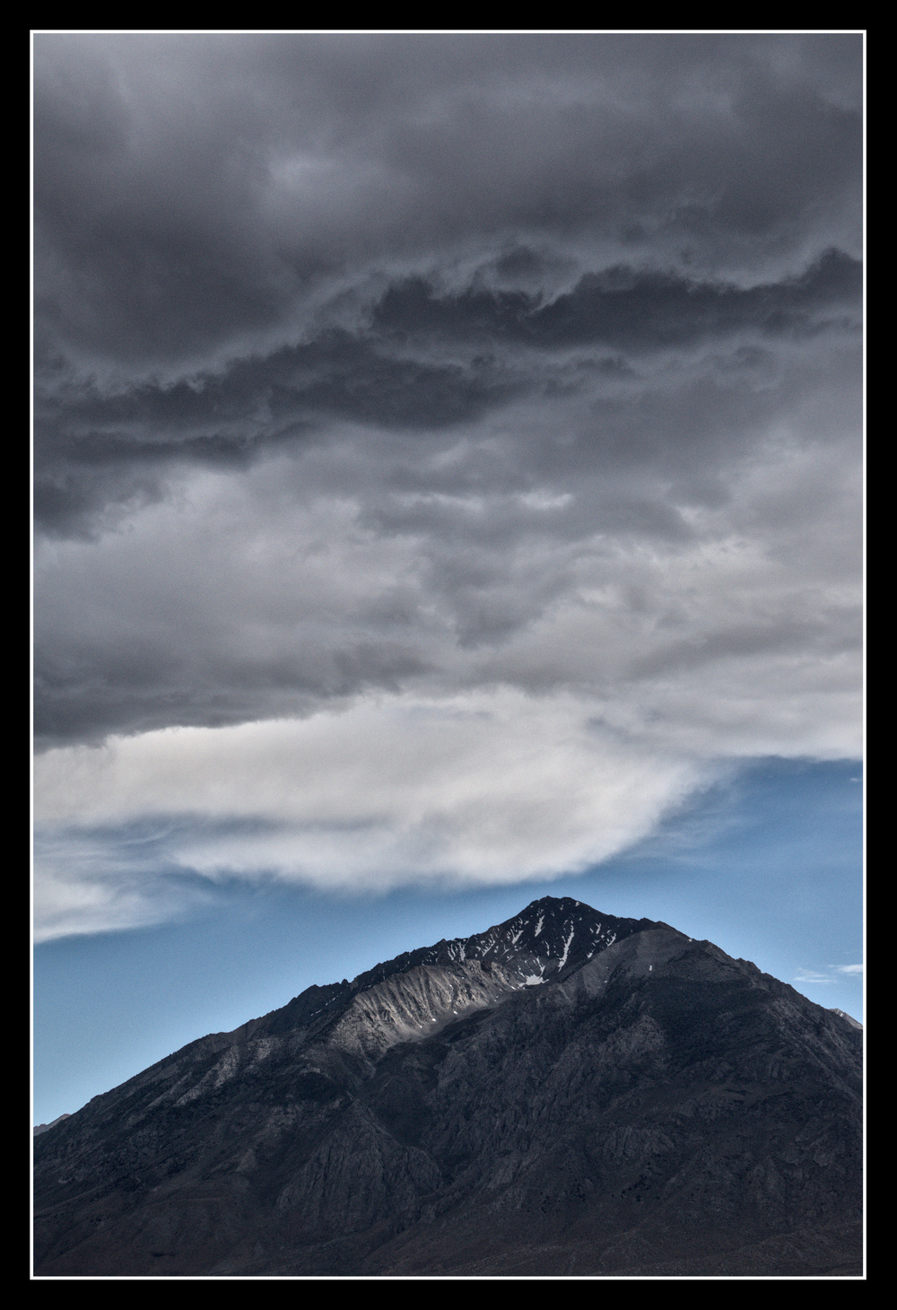 A triangular granite mountain has blue sky just above it, then clouds going from white to dark grey.
