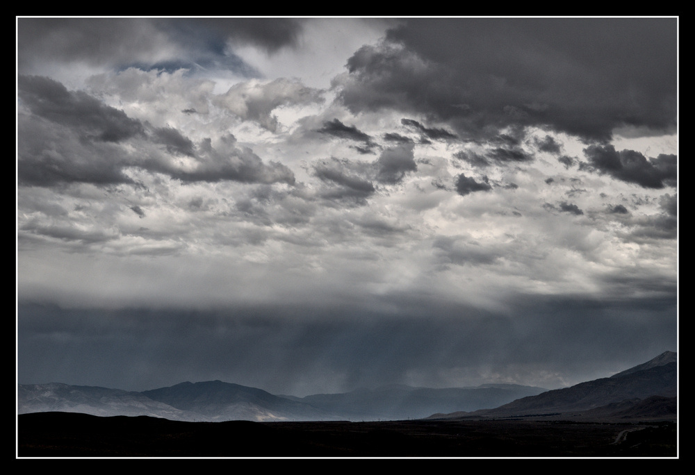 Rain falls on a valley surrounded by mountains from a dark layer of clouds near the ground.  But above a sharp line, the clouds are puffy and mostly white, with patches of blue sky.