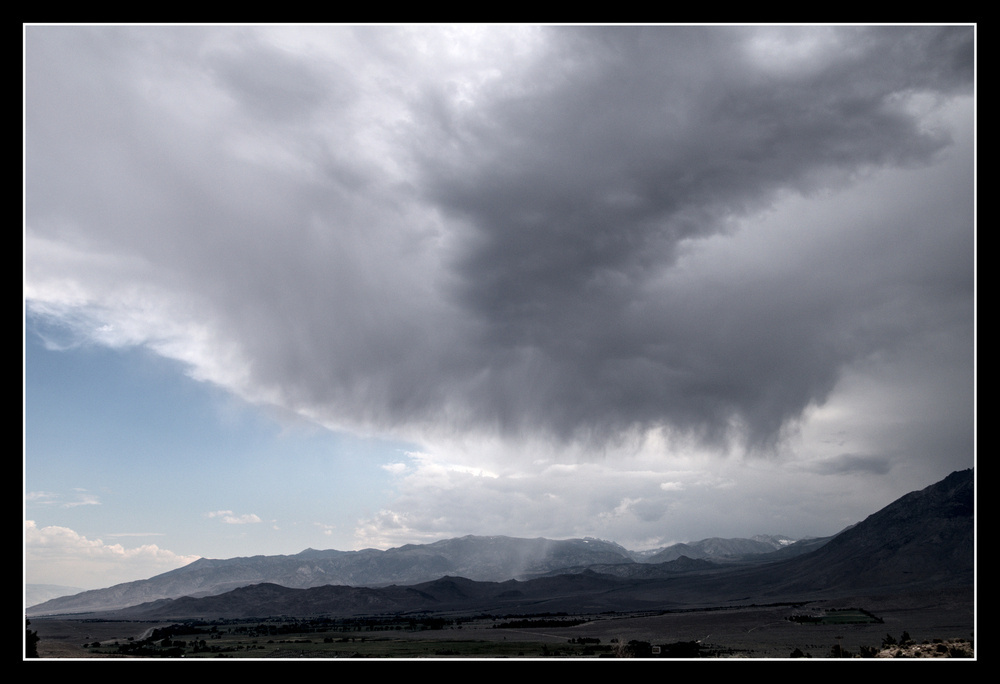 A large cloud formation over mountains is mostly light grey, turning darker at the bottom, dropping rain on the rough, rocky hills below.