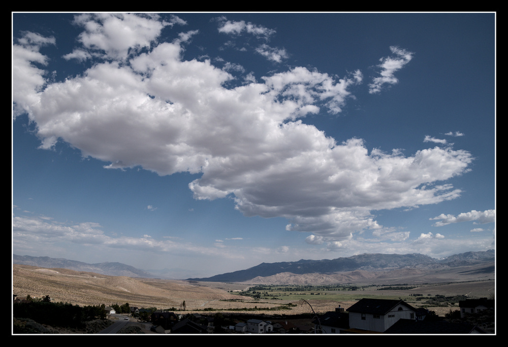 A high desert valley is brightly lit except for shadows cast by large banks of fluffy white clouds.