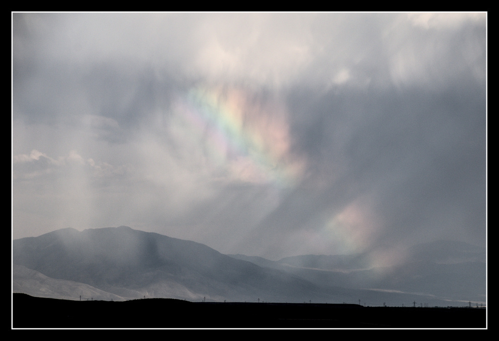 A cloudy, rainy landscape has sections of a rainbow dimly spread through it.