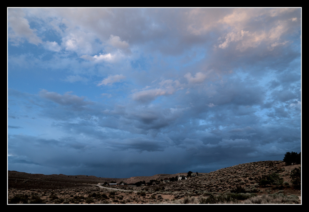 Above a valley of tan, desert plants, a large sky of clouds clouds of varying shapes and greys seem blue in the evening light.