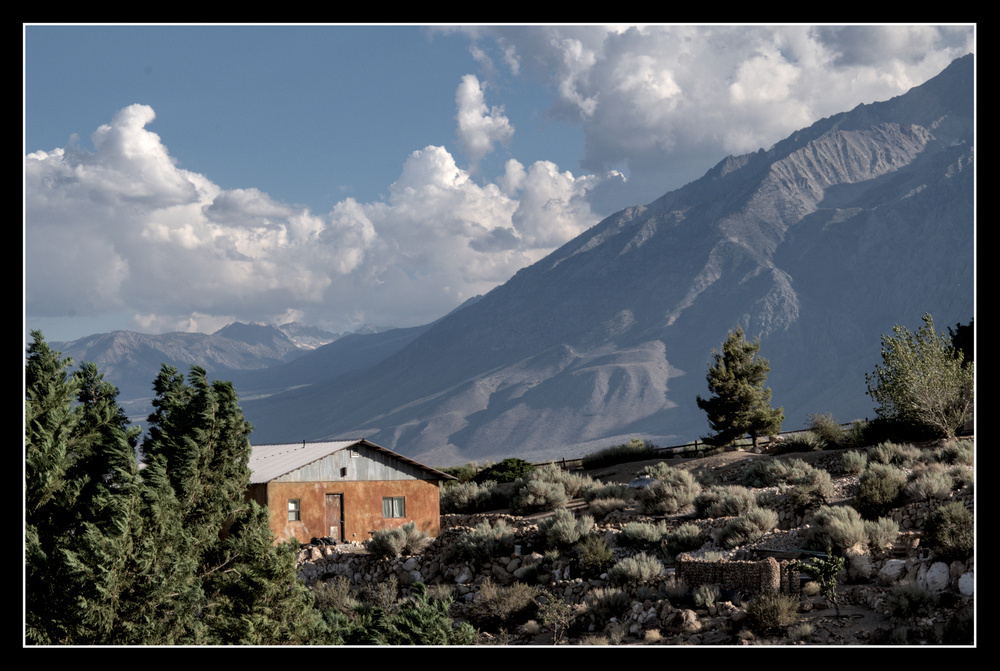 A small house with a metal roof and limestone walls sits on a hill in a valley surrounded by big mountains.
