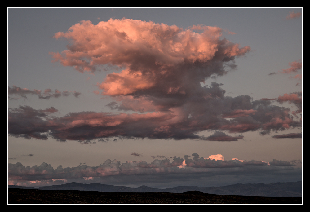 A large cloud, shaped like an anvil, lit pink by the setting sun, hovers over a long mountain range.