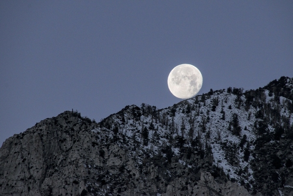 The full moon appears to just touch  a mountain ridge, dimly lit by dawn light.