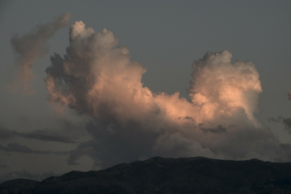 A large, double-headed cumulonimbus cloud rises above dry mountains, lit pink by the setting sun.