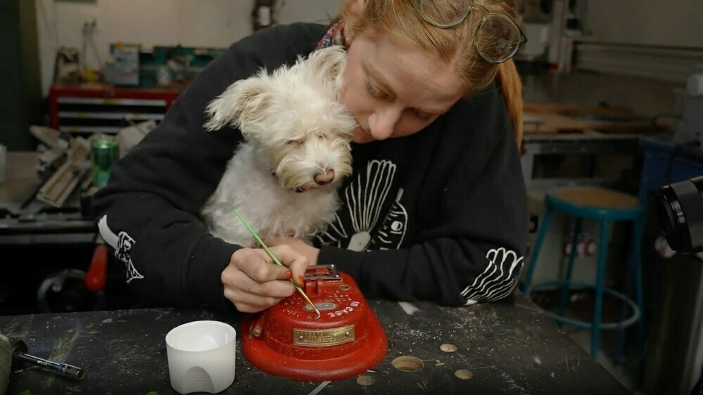 A woman sits at a desk, painting the letters of an old fire alarm. There's a dog in her lap following the paintbrush's motions.