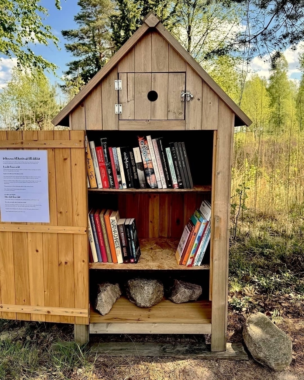 A cute little library on the beach, surrounded by trees and greenery. The door is open, showing mystery novels, Nordic noir, and some children’s books.