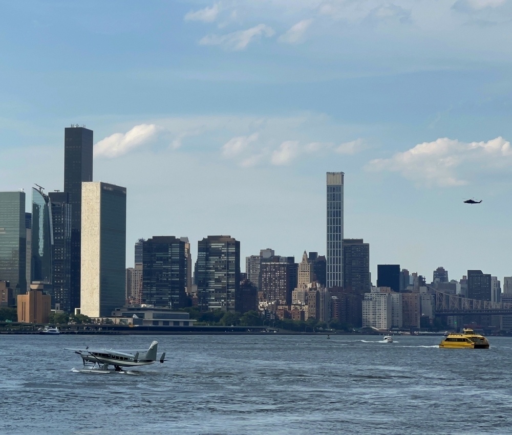 Picture of NYC's East River with skyscrapers in the background. A plane and ferry are in the water, and a helicopter is in the sky.