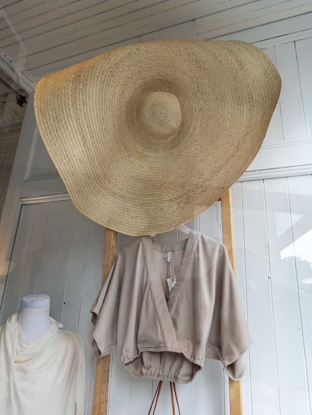 Huge brimmed woven hat hanging on wall above woman’s blouse also hanging on wall.