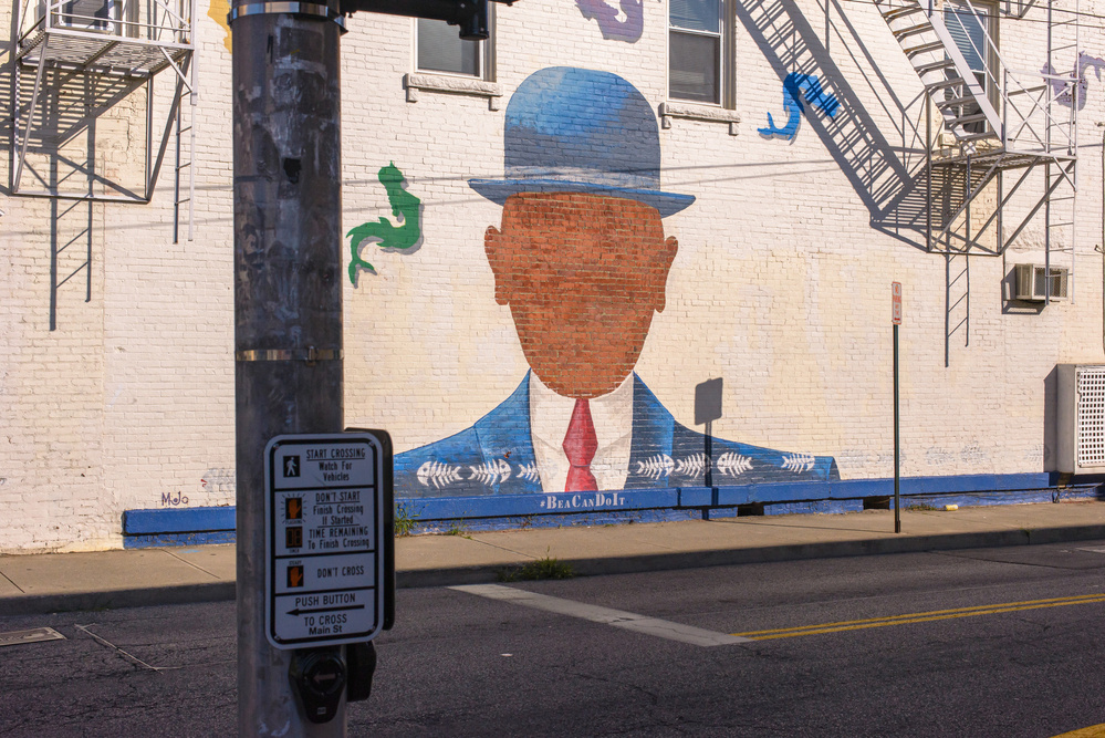 mural of head and shoulders of man with bowler hat, suit and tie, no facial features, just silhouette, fire escapes and shadows in upper left and right corners, utility pole in fore ground on left