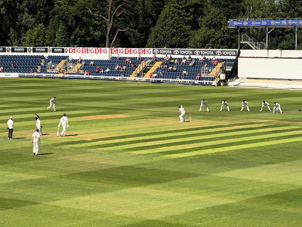 Auto-generated description: A cricket match is being played on a well-maintained field with players in white uniforms and some spectators in the stands.