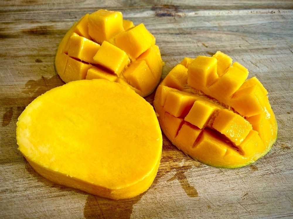 A mango sliced into thirds, two of which are hedgehog-style and one of which is the seed, all sitting on a wooden chopping board