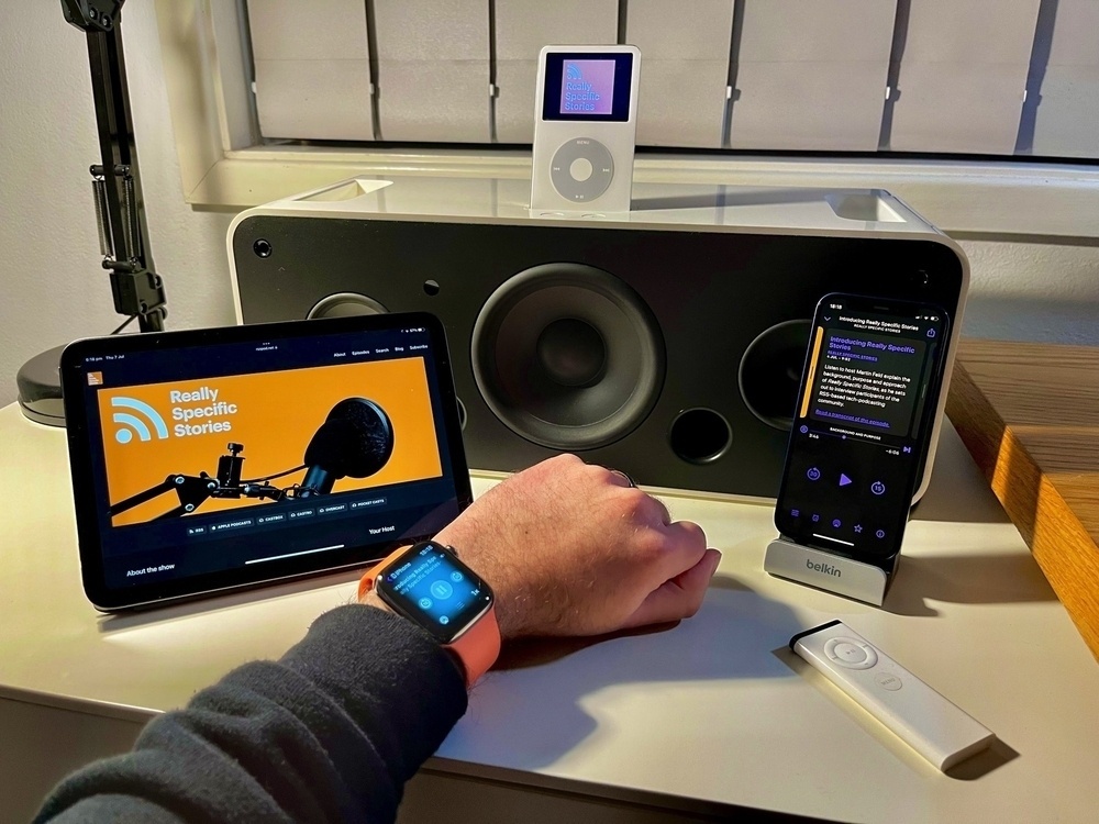 Podcast artwork and apps displayed across an Apple Watch, iPad mini (6th gen.), iPhone 12 mini and white iPod Video docked in an iPod Hi-Fi